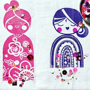 May include: Two fabric prints of stylised figures with rainbow arches, decorated with pink, white and gold sequins. The figures are on a white background.
