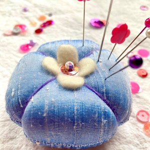 May include: A blue fabric pincushion shaped like a flower with a white felt center and pink flower accents. The pincushion has several pins with pink flower heads sticking out of it.