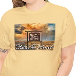 Peut inclure: Un t-shirt jaune clair avec une image panoramique d'un panneau de bienvenue du Colorado sous un coucher de soleil coloré. Le panneau indique "Welcome to Colorful Colorado" avec "State of Mind" en dessous en écriture cursive. Le t-shirt est en matière douce.