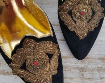 May include: A pair of black suede heeled shoes with gold embroidery and red jewels. The shoes have a pointed toe and a gold leather lining. The size is marked as "A 4080" on the inside of the shoe.