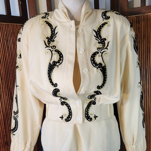 May include: Cream-colored two-piece suit with black and gold embroidered detailing. The jacket has a button-up front and long sleeves. The skirt is a straight, knee-length style. The suit is displayed on a mannequin.