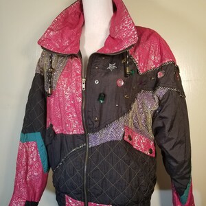 May include: A vintage, quilted jacket with a vibrant design. The jacket features a mix of black, pink, and teal panels, embellished with decorative elements. It has a high collar, a zip-up front, and a quilted pattern on the black sections.