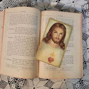 May include: A vintage religious bookmark featuring a close-up image of Jesus Christ with a red heart and flames. The bookmark is printed on cardstock and has a white border.