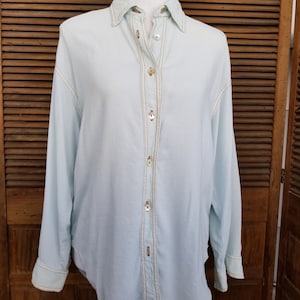 May include: A light blue long-sleeved button-down shirt with gold stitching detail down the front.