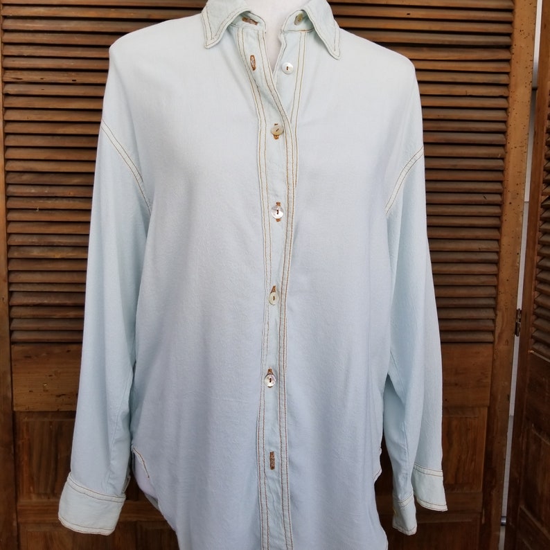 May include: A light blue long-sleeved button-down shirt with gold stitching detail down the front.