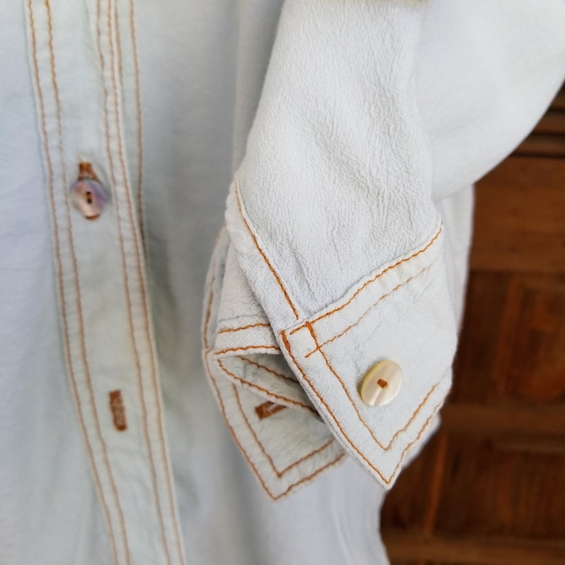 May include: A light blue button-up shirt with contrasting orange stitching detail. The shirt has a button closure and a long sleeve with a button cuff.