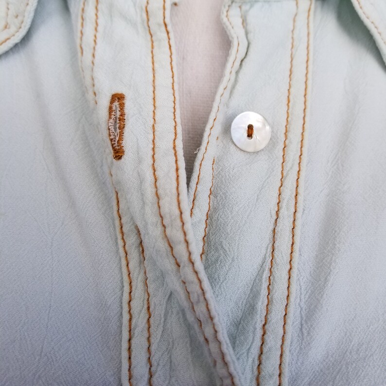 May include: A light blue button-up shirt with a white button and brown stitching.