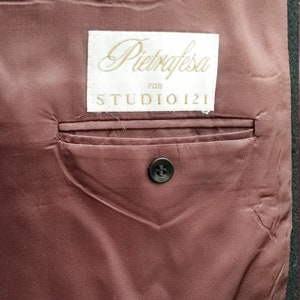 May include: Close-up of the interior of a jacket, featuring a dark burgundy lining. A small pocket is visible, along with a black button. A label reads "Pietrafesa for STUDIO 121".