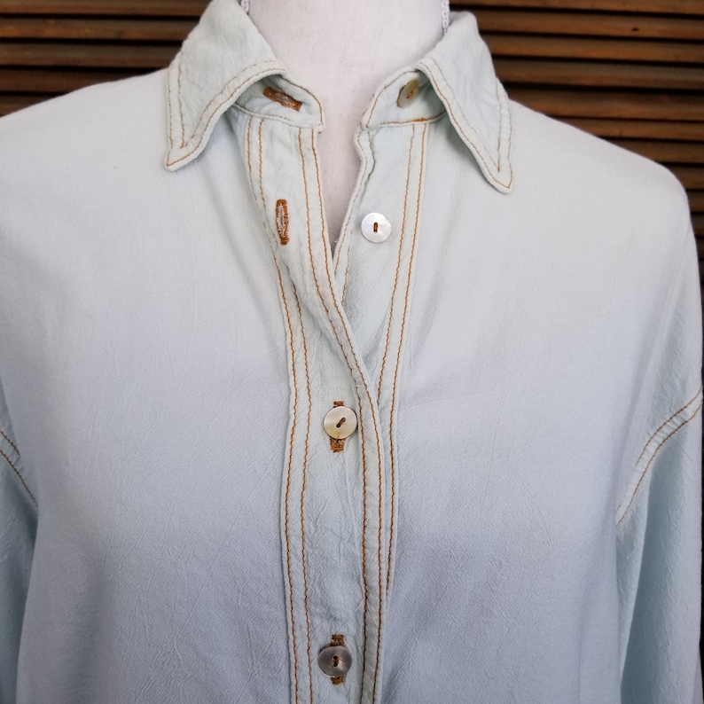 May include: A light blue button-down shirt with brown stitching and mother-of-pearl buttons.