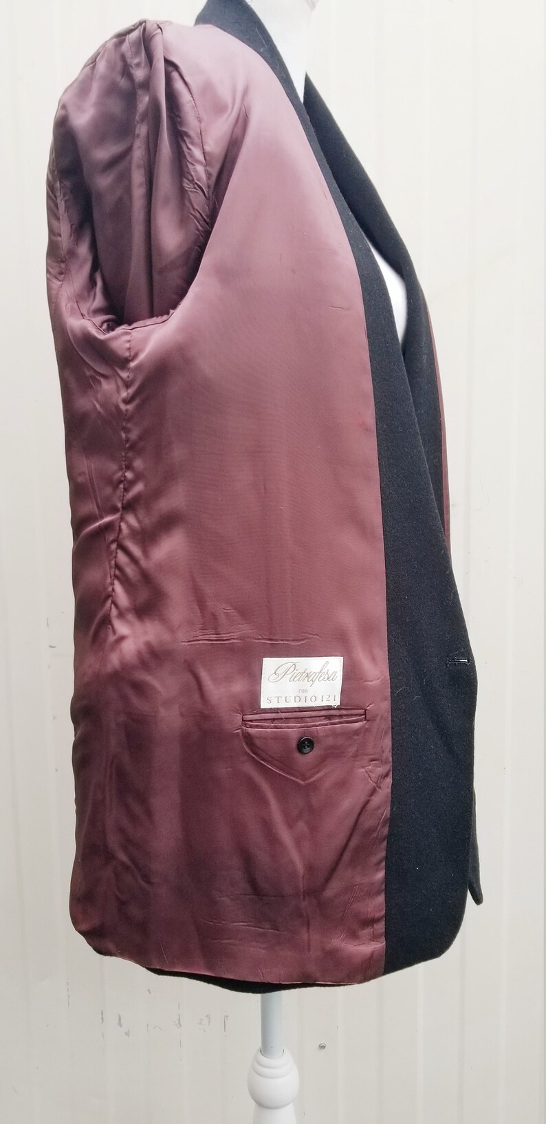 May include: Black blazer with a burgundy satin lining. The interior features a pocket with a button closure and a label that reads "Pietrafesa STUDIO 121". The blazer is displayed on a white mannequin.