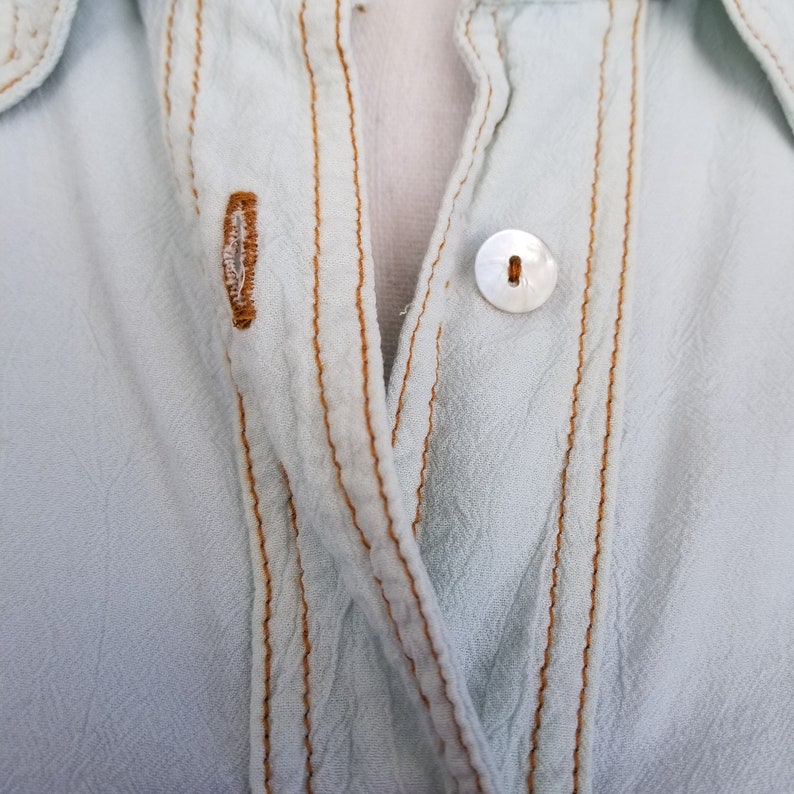 May include: A light blue button-up shirt with a white button and brown stitching around the front placket.