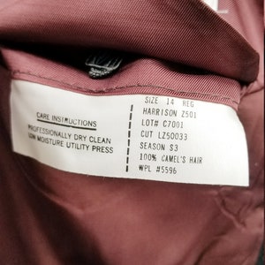May include: Close-up of the inside of a burgundy-colored garment, revealing a label with care instructions and details like "SIZE 14 REG" and "100% CAMEL'S HAIR". The label also includes the text "HARRISON Z501".