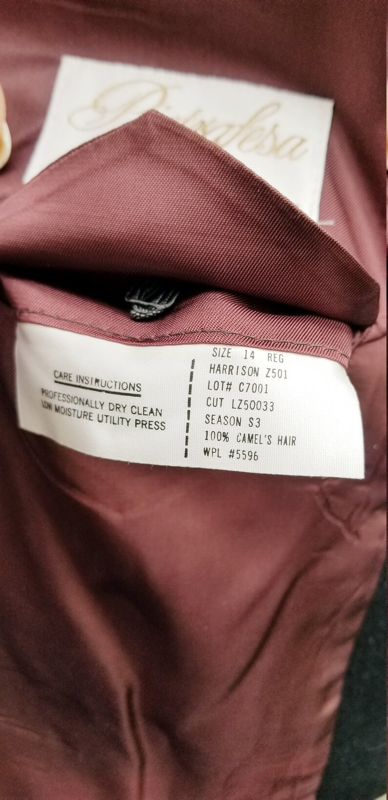 May include: Close-up of the inside of a burgundy-colored garment, revealing a label with care instructions and details like "SIZE 14 REG" and "100% CAMEL'S HAIR". The label also includes the text "HARRISON Z501".