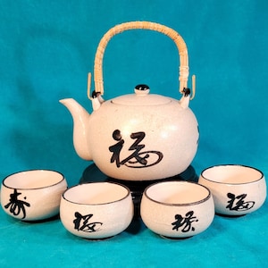 May include: A white ceramic teapot with a bamboo handle and black calligraphy. The teapot is surrounded by four matching white ceramic teacups with black calligraphy. The teacups and teapot have a speckled finish.
