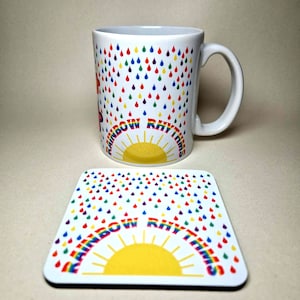 May include: White ceramic mug featuring a rainbow design and the text "Rainbow Rhythms". The mug has a yellow sun and colourful raindrops. A matching coaster with the same design is also shown.