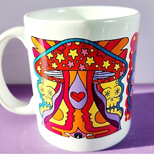 May include: White ceramic mug with a colorful psychedelic design featuring a mushroom with stars, a heart, and two skulls. The design is in shades of red, orange, yellow, purple, and blue.
