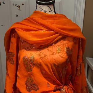 May include: An orange dress with gold floral patterns and a matching scarf draped over a mannequin. The dress is gathered at the waist and has long sleeves. The mannequin has a black top and a white base.