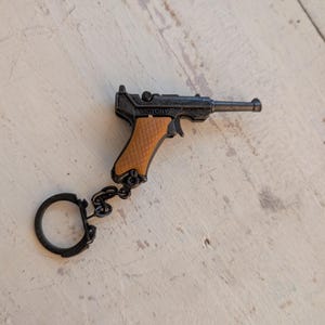 Op de afbeelding: Een miniatuur zwart en bruin pistool sleutelhanger met een bruin handvat en het woord "VICTORY" aan de zijkant. De sleutelhanger is bevestigd aan een zwarte ketting en ring. Het pistool is van metaal.