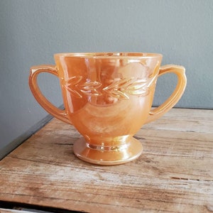 Pode incluir: Um cremoso ou açucareiro vintage de vidro cor de pêssego com duas alças e um design de folhas. A tigela tem um acabamento iridescente.