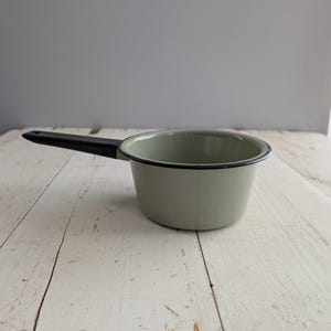 May include: A small, sage green saucepan with a black handle and rim. The enamel pot sits on a distressed, white wooden surface. The handle is angled to the left, suggesting it is ready for use in the kitchen.