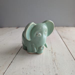 Op de afbeelding: Een kleine, lichtgroene keramische olifantenfiguur. De olifant heeft grote oren, een slurf en kleine slagtanden. Het beeldje staat op een wit houten oppervlak. De olifant is een decoratief item.