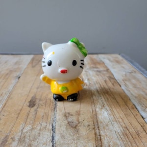 May include: A small white ceramic figurine of Hello Kitty wearing a yellow dress with a green bow. The figurine has black eyes and a red nose.