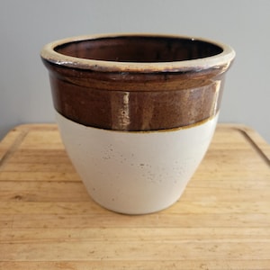 以下が含まれることがあります： 上部に茶色の釉薬をかけた白い陶器の壺。壺は木製の表面の上に置かれています。