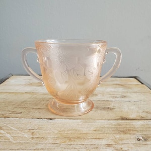 Könnte beinhalten: Eine antike Zuckerdose aus rosa Glas mit zwei Henkeln und einem Blumendesign.