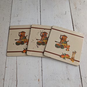 May include: Three vintage square trivets with a cream background and a brown border. Each trivet features a colorful illustration of a teapot, fruit, and utensils. The trivets are made of a durable material and are ideal for protecting surfaces from heat.