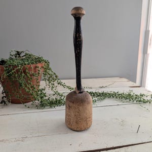 Puede incluir: Un mortero de madera vintage con un mango oscuro y una cabeza redondeada. El mortero tiene una base cilíndrica y una apariencia desgastada y rústica, lo que sugiere que es una herramienta de cocina antigua. El mango es negro con desgaste visible.