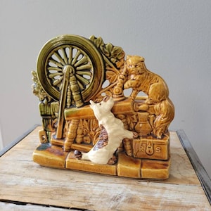 May include: A ceramic planter shaped like a scene with two cats, one sitting by a spinning wheel and the other standing on a brick path. The planter is brown and green with a white cat and the words "R.S." on the bottom.