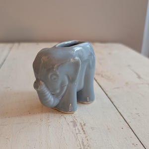 Può includere: Piccola statuetta di elefante in ceramica azzurra con la parte superiore scavata. L'elefante ha una proboscide, orecchie e quattro zampe. La statuetta è smaltata e ha una finitura lucida. L'elefante è un oggetto decorativo.