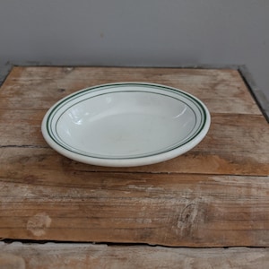 May include: White oval serving bowl with a green double stripe around the rim. The bowl is set on a rustic wooden surface. This vintage-style bowl is suitable for serving side dishes or appetizers.