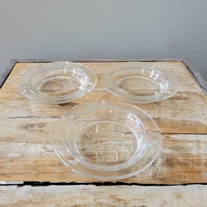 Peut inclure: Trois sous-verres en verre transparent avec un motif en étoile au centre. Les sous-verres sont sur une surface en bois.