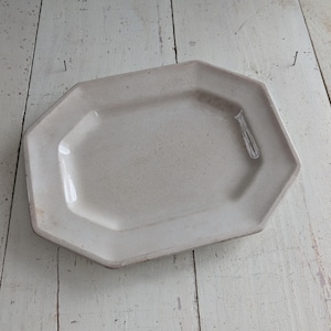 以下が含まれることがあります： オフホワイトの八角形のサービングプラッター。セラミック製の皿は長方形の中央部とわずかに盛り上がった縁があります。表面には使用感があり、アンティークな魅力を添えています。プラッターは、白く風化した木製の表面に置かれています。