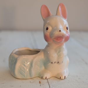 Op de afbeelding: Een vintage keramische konijnenplanter. Het konijn is wit met roze oren en wangen, en lichtblauwe bladdetails. De planter is ontworpen in een zittende positie met een kleine opening voor planten.