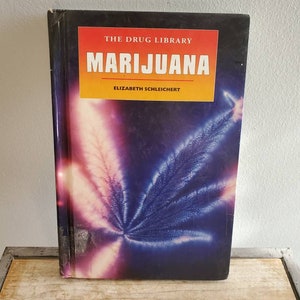 Può includere: Un libro intitolato "Marijuana" della serie "The Drug Library". La copertina presenta una foglia di cannabis stilizzata in tonalità viola, rosa e blu su uno sfondo scuro. È visibile anche il nome dell'autore, Elizabeth Schleiert.