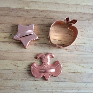 Puede incluir: Tres cortadores de galletas de cobre en forma de estrella, manzana y flor de lis.
