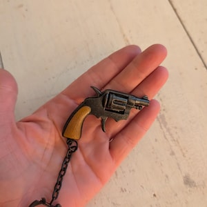 Könnte beinhalten: Miniatur-Revolver-Schlüsselanhänger in Schwarz und Gold mit Holzoptik-Griff. Der Schlüsselanhänger hat eine kleine Kette und einen runden Schlüsselring. Der Revolver ist eine detaillierte Nachbildung einer klassischen Schusswaffe.