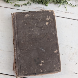 Puede incluir: Un antiguo y desgastado diccionario Webster's New Handy Dictionary con una cubierta marrón oscuro. La cubierta del libro está desgastada con desgaste visible, y el título está impreso en un diseño circular. El libro está sobre una superficie de madera blanca.