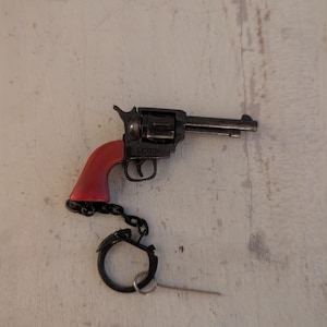 Op de afbeelding: Een kleine zwarte speelgoedrevolver met een rode handgreep, bevestigd aan een sleutelhanger. Het woord "VICTORY" is zichtbaar op het frame van het pistool. De sleutelhanger is zwart en bevat een ring en een metalen pin.