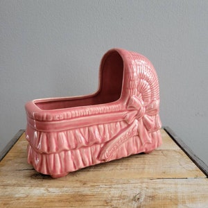 Puede incluir: Maceta de cerámica rosa en forma de cuna de bebé con una cinta blanca y la palabra "Felicidades" en el lateral.