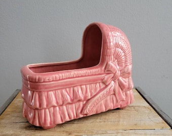 May include: Pink ceramic baby crib planter with a white ribbon and the word "Congratulations" on the side.