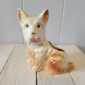 May include: A ceramic dog planter with a white and brown glaze. The dog is sitting with its head tilted and has a pink nose and brown eyes.