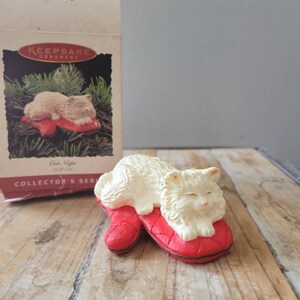 May include: White porcelain cat figurine ornament sleeping on a red mitten. The ornament is part of the Hallmark Keepsake Ornament Collector's Series.