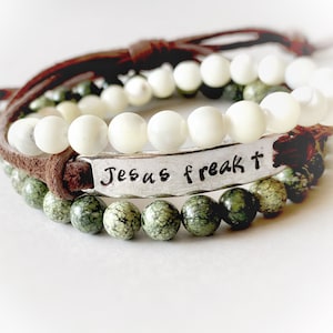 May include: A set of three bracelets with a brown leather cord, white beads, and green beads. The leather cord has a silver charm with the text "Jesus freak t".