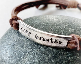 May include: A brown leather bracelet with a silver bar that reads "deep breaths" in a handwritten font. The bracelet is resting on a gray stone. The bracelet is adjustable.
