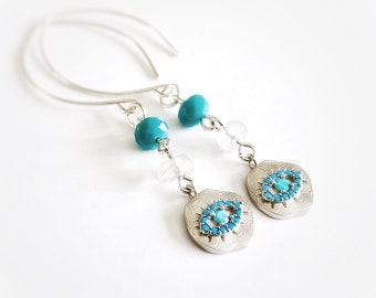 May include: Silver-tone dangle earrings featuring turquoise-colored beads and eye-shaped pendants. The pendants have a textured surface and are adorned with small turquoise stones. The earrings have a simple, elegant design.