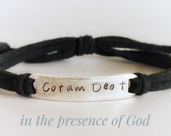 May include: A black leather bracelet with a silver charm that reads "Coram Deo +" in cursive. The charm is a rectangular shape with rounded edges. The bracelet is tied with a knot.