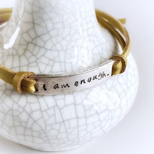 May include: A mustard yellow leather bracelet with a silver charm that reads "I am enough."